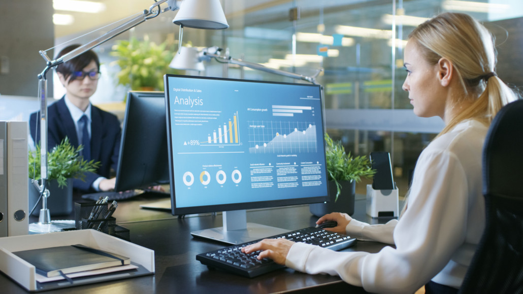 Financier Works on a Personal Computer Showing Statistics, Graphs and Charts. In the Background His Coworker and Creative Office.