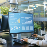 Financier Works on a Personal Computer Showing Statistics, Graphs and Charts. In the Background His Coworker and Creative Office.